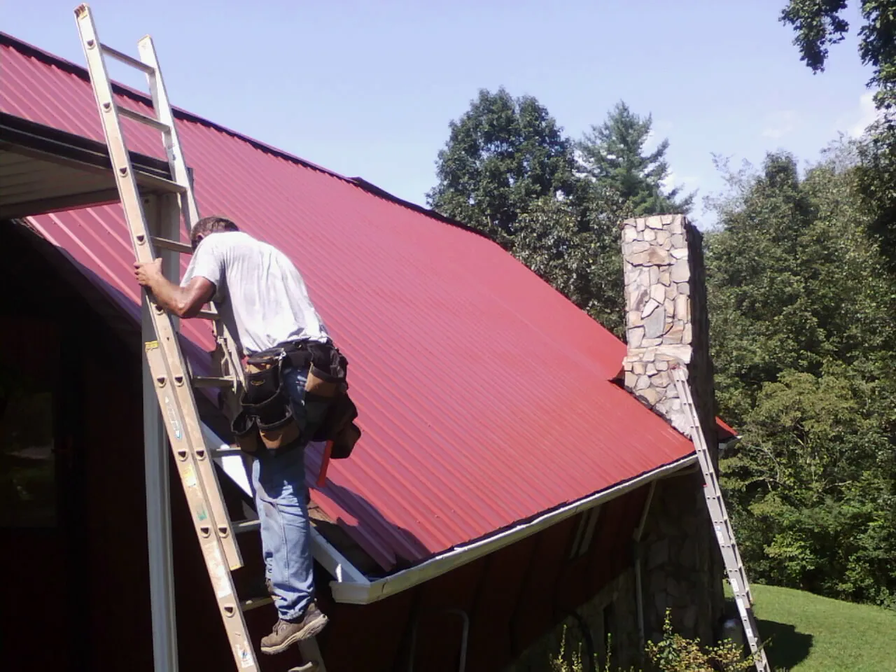 Roofer on a ladder beside a red metal roof and stone chimney — Soffit & Fascia Repair by Roofing Brothers in Saltese